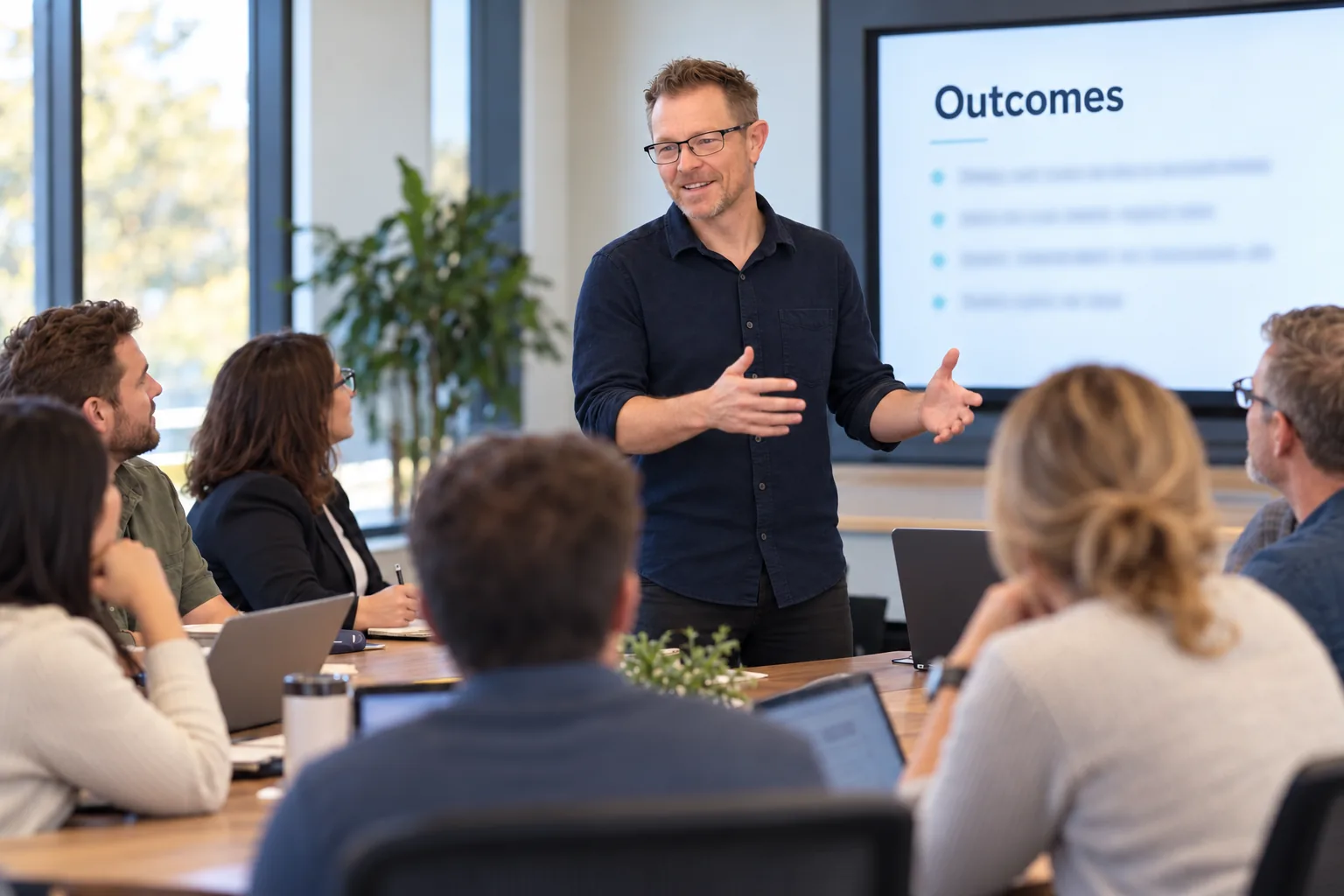 Adam Michaelson standing at the front of a conference room, gesturing as he trains a seated team, with a slide titled Outcomes on the screen behind him