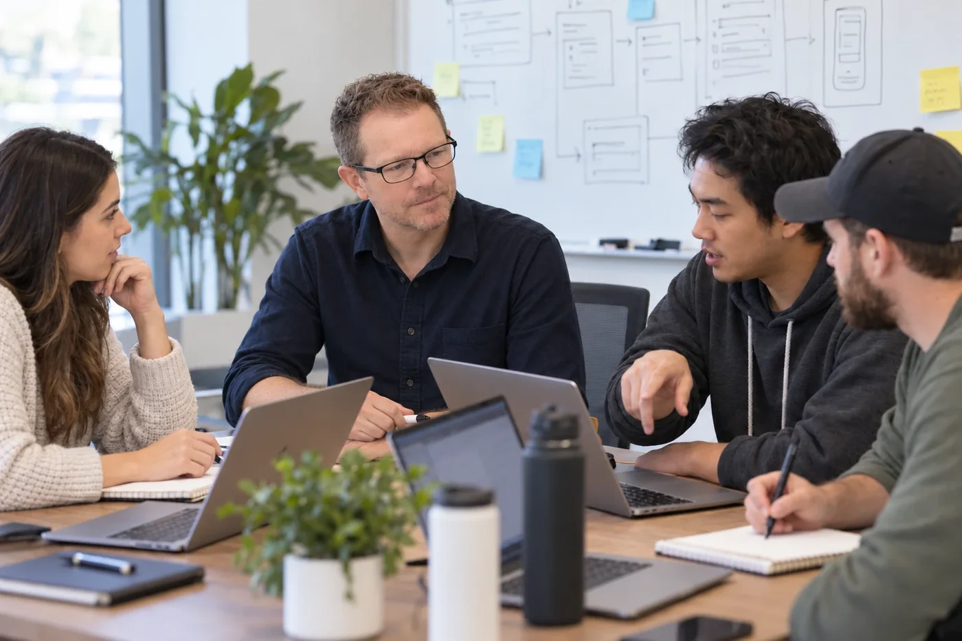 A product team of four works around a table with laptops and notebooks, with app wireframes and sticky notes on the whiteboard behind them